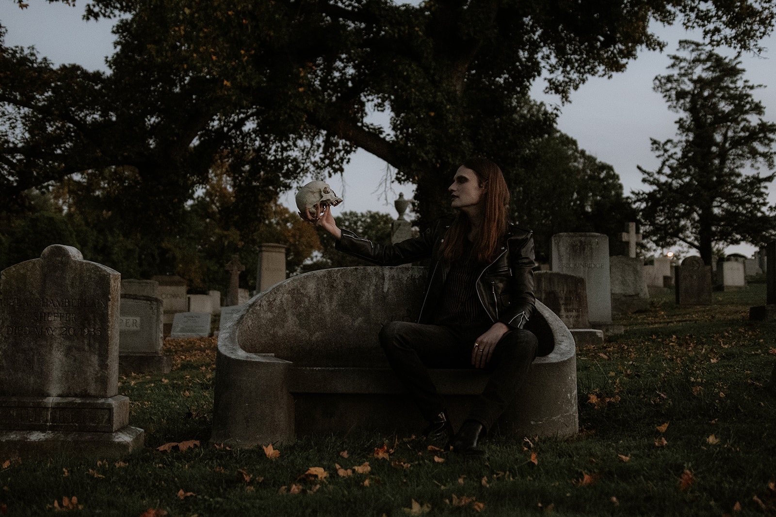 Hunter sits in a graveyard, holding up a fake skull to gaze at. He's wearing a leather jacket and all-black basics. His long brown hair is down.