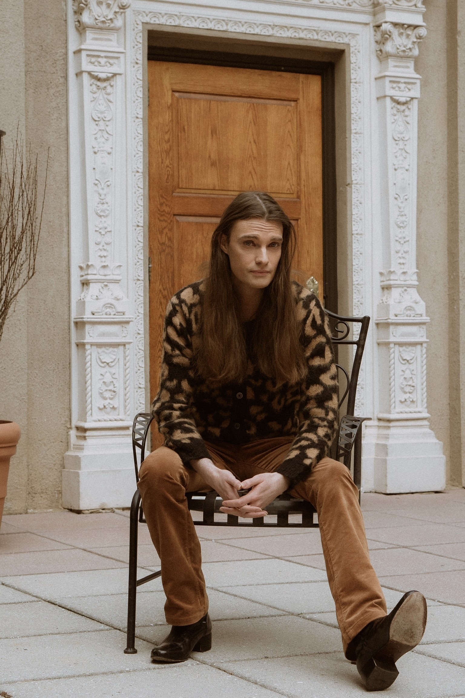 Hunter sits in a courtyard wearing brown velvet flare trousers, heeled black leather boots, and a leopard print sweater. His long brown hair is down.